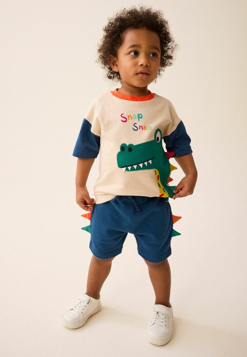 Toddler in a cream shirt with a green dinosaur graphic and blue shorts with colorful dinosaur spikes, wearing white sneakers and standing.