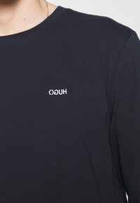 Marineblaues Langarmshirt aus weicher Baumwolle, mit einem kleinen weißen gestickten Logo auf der Brust. Einfaches Rundhalsdesign.