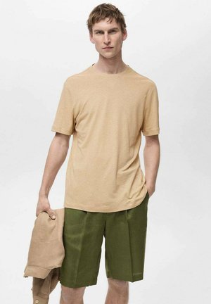 T-shirt in cotone di un leggero beige con colletto a girocollo, abbinato a pantaloni corti verde oliva. Il modello tiene una giacca beige in una mano, in piedi contro uno sfondo neutro.