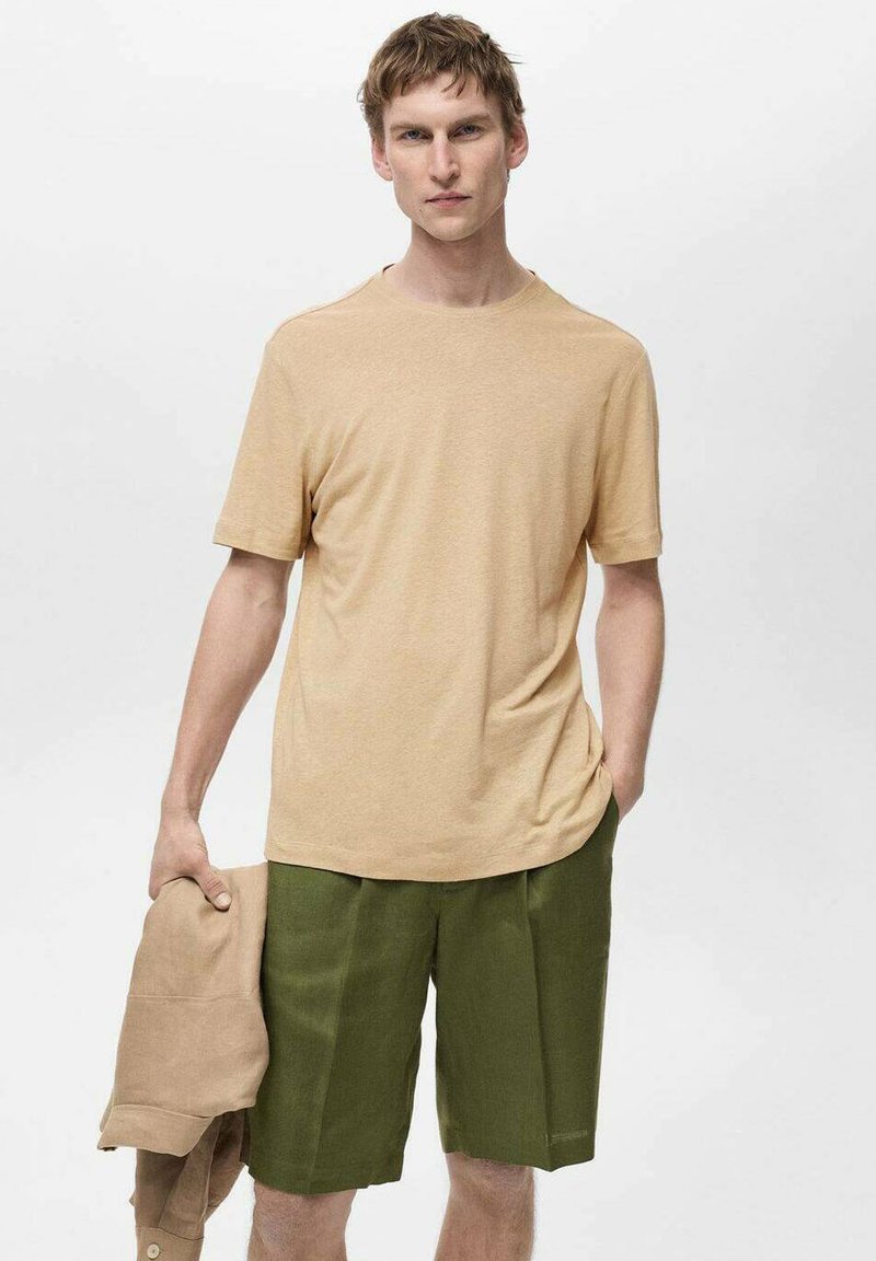 Vala beige tekstiilit-paita, jossa on pyöreä kaula-aukko, yhdistettyinä oliivinvihreisiin shortseihin. Malli pitää beigeä takkia yhdessä kädessä seisten neutraalia taustaa vasten.