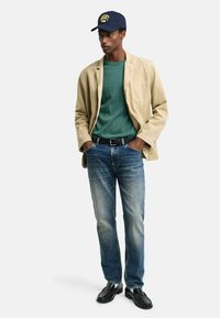 Beige Blazer, grüner Rundhalsausschnitt-Pullover, blaue Jeans und schwarze Slipper. Marineblaues Cap mit gelbem Logo. Slim Fit, lässiger Stil.