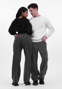 Par i afslappet tøj; den ene i en sort cropped hoodie, den anden i en hvid sweatshirt. Begge iført grå vide jeans og sorte sko.
