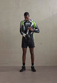 Homme portant un équipement de boxe noir et vert ajuste des bandages blancs contre un fond de mur gris uni.