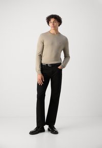 Beige gestricktes Langarmshirt mit einem kleinen roten Logo, kombiniert mit schwarzen Hosen und schwarzen Schnürschuhen auf einem weißen Hintergrund.