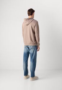 Man in een beige hoodie, blauwe jeans en lichte sneakers, die met zijn rug naar de camera staat tegen een effen witte achtergrond.