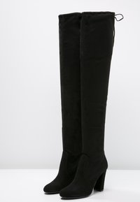 Botas negras de ante hasta la rodilla con punteras redondeadas y tacones gruesos, de pie sobre una superficie blanca contra un fondo gris claro.