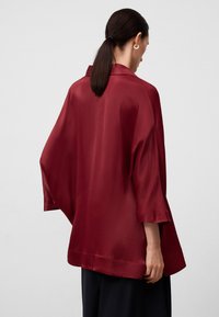 Bordowa satynowa kurtka w stylu kimono z szerokimi rękawami, gładką fakturą i luźnym krojem, przedstawiona z tyłu na jasnym tle.