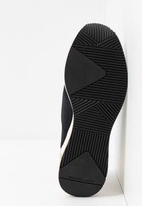 Semelle de basket noire avec un motif de grip texturé et le nom de marque "MICHAEL KORS", reposant verticalement contre une surface blanche.