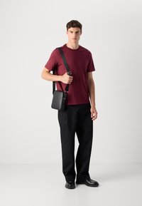 Man in een bordeauxrode t-shirt, zwarte broek en zwarte schoenen, die een zwarte schoudertas met een brede strap over zijn schouder houdt.