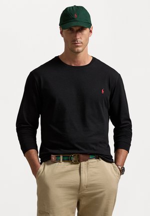 Zwarte lange mouwen shirt met een klein rood logo, beige broek, bruine riem met groene en blauwe strepen, en een groene pet met rood logo.