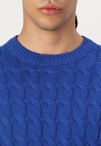 Blauer Strickpullover mit Zopfmuster und einem gerippten Rundhalsausschnitt, der auf glattem Stoff eine strukturierte Optik zeigt.