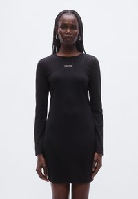 Juoda ilgomis rankovėmis suknelė iš lygaus audinio, suapvalintu kaklu ir aptempta silueto linija, krūtinės srityje subtiliai pažymėta Calvin Klein logotipu.