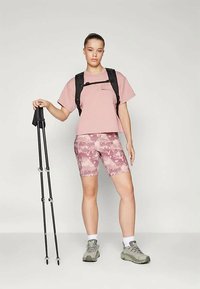 Light pink cropped t-shirt with a chest pocket, paired with pink camo-patterned bike shorts and gray hiking shoes. Black backpack and trekking poles.