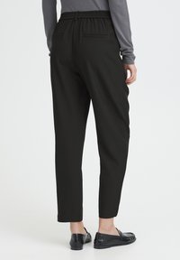 Pantalon noir fuselé avec une taille élastique et des poches arrière, associé à des mocassins noirs, mettant en avant un design minimaliste et une texture lisse.