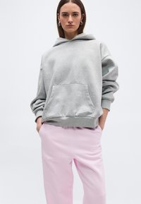 Νεαρή γυναίκα με ίσια καστανά μαλλιά που φοράει ασημένια σκουλαρίκια κρίκους, ανοιχτογκρι φαρδιά μπλούζα με κουκούλα και ανοιχτορόζ παντελόνι με τα χέρια στις τσέπες.