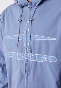 Giacca azzurra chiara con logo PXLD bianco e design a stella sul petto, dotata di cerniera e coulisse con terminali metallici.