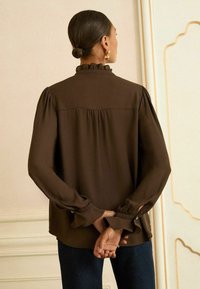 Una donna dalla pelle scura e capelli raccolti in un bun indossa una blusa marrone a maniche lunghe con colletto e polsini a balze, con le mani incrociate dietro la schiena, di fronte a un muro.