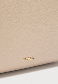 Μπεζ τσάντα από υφή δέρματος με ανάγλυφο χρυσό λογότυπο "FURLA". Ο σχεδιασμός είναι μινιμαλιστικός με ομαλές άκρες και καθαρές γραμμές.