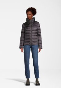Doudoune grise avec un col haut et fermeture éclair, dotée d'un design matelassé horizontal, associée à un jean bleu et des bottines.