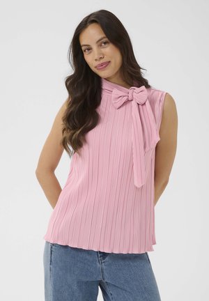 Žena s dlhými tmavými vlasmi, ktorá má na sebe ružový plisovaný bezrukávový top s mašľou na krku a modré rifle, stojaca pred jednoduchým pozadím.