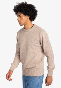 Man met krullend zwart haar, gekleed in een beige gebreide trui en een blauwe spijkerbroek, staande in profiel tegen een witte achtergrond.