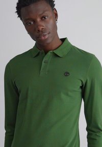 Camisa polo de manga longa verde com um acabamento texturizado e colarinho. Apresenta um pequeno logótipo preto na parte esquerda do peito.