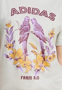 Bela majica s kratkimi rokavi, ki prikazuje grafični design dveh vijoličnih ptic, obdanih z rumenimi cvetovi, z napisom "ADIDAS" in "FARM RIO" v kostanjevi barvi.