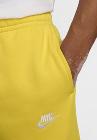 Persona con vitiligo, mano en el bolsillo, usando pantalones de chándal amarillos de Nike con logo blanco y cintura elástica, frente a un fondo blanco.