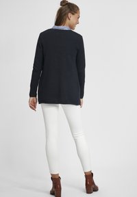 Pull en maille texturé bleu marine avec des manches longues et un col rayé, associé à des leggings blancs et des bottines brunes, vu de dos.