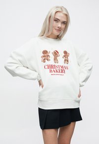 Balta svītra ar trim piparkūku figūrām un sarkano tekstu "Ziemassvētku maiznīca" ar svinīgu dizainu. Garās piedurknes, ērta piegriezuma.