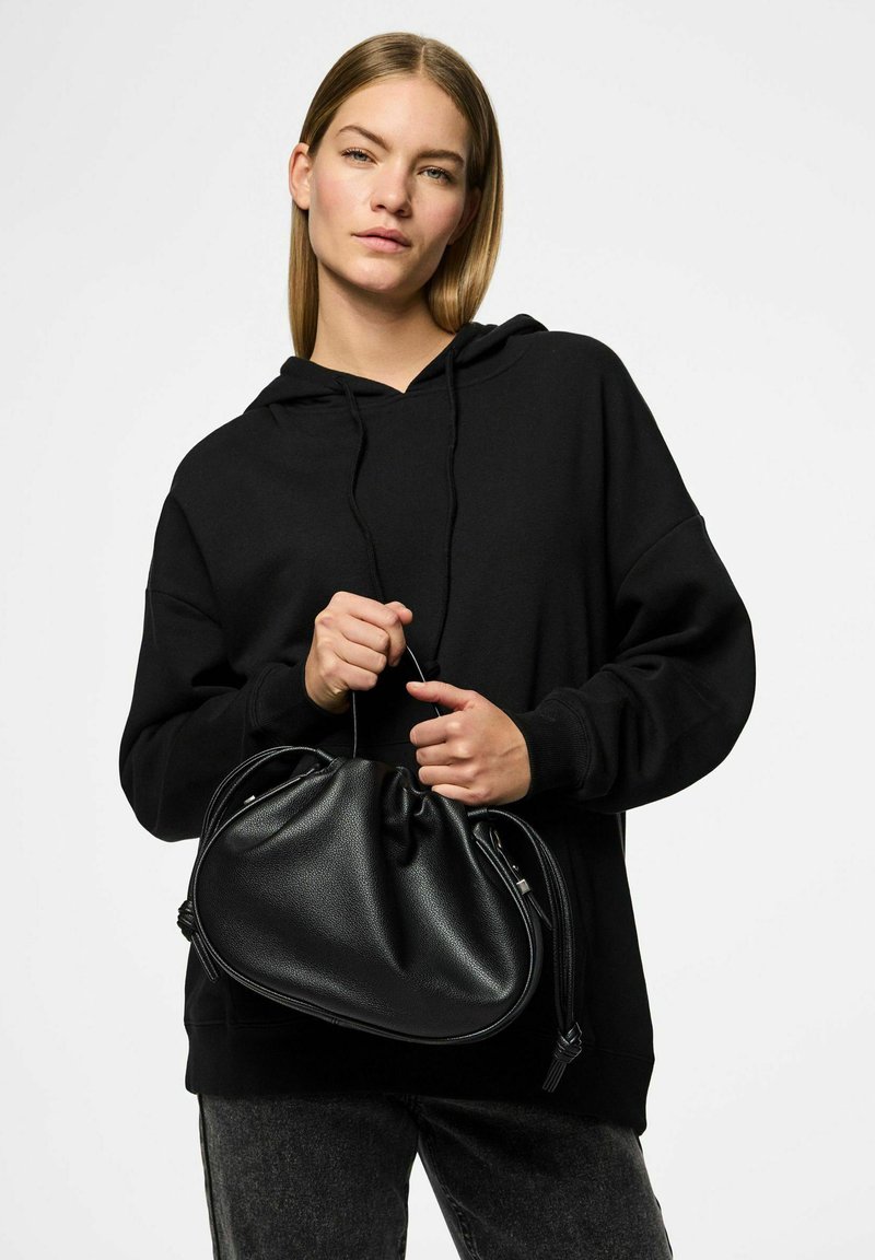 Borsa a mano nera in pelle con una superficie morbida e testurizzata, forma arrotondata, apertura cinchata e leggeri dettagli metallici, tenuta tra le mani di una persona.