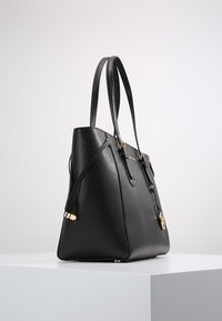 Bolso tote de cuero negro con herrajes dorados, doble asas para el hombro y un charm decorativo colgante sobre un pedestal blanco contra un fondo liso.