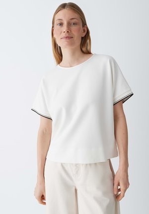 Donna con capelli chiari che indossa una maglietta bianca a maniche corte con bordi neri sulle maniche e pantaloni color crema su uno sfondo chiaro uniforme.