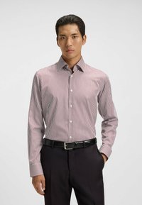 Homme portant une chemise à manches longues rayée rouge et blanche, rentrée dans un pantalon noir avec une ceinture noire, se tenant contre un fond uni.