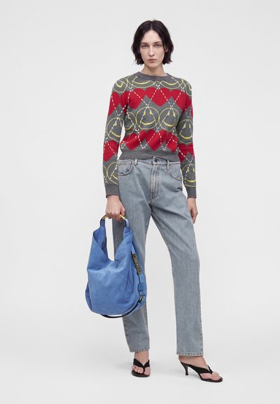 Femme portant un pull gris avec des cœurs rouges et des visages jaunes souriants, jean bleu clair, talons noirs ouverts à l’avant, tenant un grand sac à main bleu.