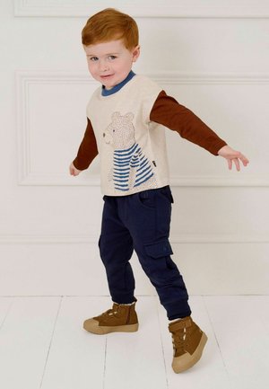 Kinderoutfit mit einem beigen Langarmshirt mit Bärendesign und einer marineblauen Cargohose. Braune Stiefel runden den Look ab.