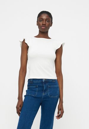 Femme debout devant un fond blanc, portant un chemisier sans manches blanc avec des épaules à volants et un jean bleu taille haute.