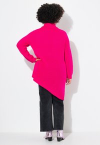 Rosa strukturierter Pullover mit hohem Kragen und asymmetrischem Saum, kombiniert mit dunklen, weit geschnittenen Hosen und glänzenden silbernen Ankle Boots.