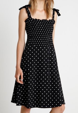 Schwarzes Kleid mit weißen Tupfen, einem gerafften Oberteil und Schulterbändern, das einen ausgestellten Rock besitzt. Glatter Stoff, gleichmäßig verteilte weiße Tupfen.