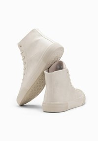 Zapatillas altas en material blanco roto, con una suela de goma texturizada, diseño de cordones y parte superior lisa con un sutil detalle en las costuras.