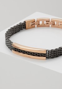 Zwarte gevlochten metalen armband met twee gepolijste rosegouden stangen met gegraveerd merk. Het ontwerp van de sluiting bevordert een veilige sluiting.