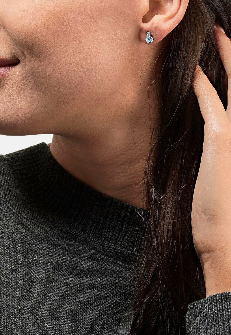 Silberne Ohrstecker mit Aquamarinsteinen, runde Form und strukturiertes Design, getragen von einer Person mit dunklen Haaren, die einen dunklen Pullover trägt.