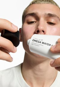 Hvid cylindrisk flaske mærket "OMEGA WATER CREAM" holdt i den ene hånd, sort låg i den anden, med en enkel baggrund og fokus på tekstur.