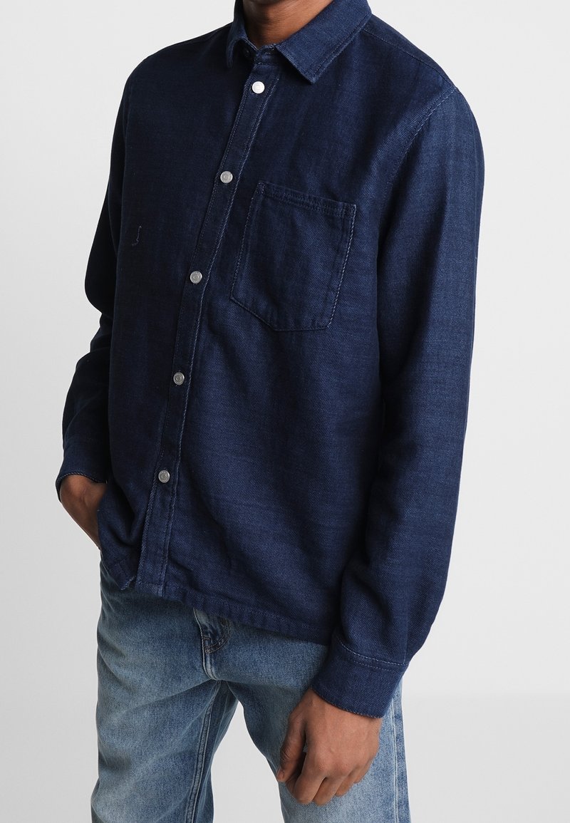 Chemise en denim bleu foncé à manches longues, boutonnée sur le devant, avec une poche poitrine unique et un tissu texturé. Portée avec un jean bleu clair.