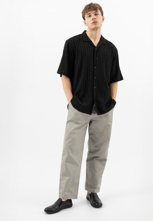 Jonge man staand met handen in zijn zakken, gekleed in een zwart kortärmelig overhemd met knopen, lichtgrijze broek en zwarte instapschoenen.