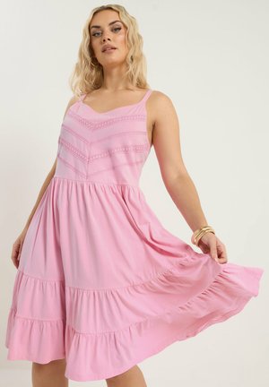 Kvinde iført en ærmeløs lyserød lagdelt kjole med blonder, der let holder kjolen ud, accessoriseret med guldringearmbånd og armbånd.