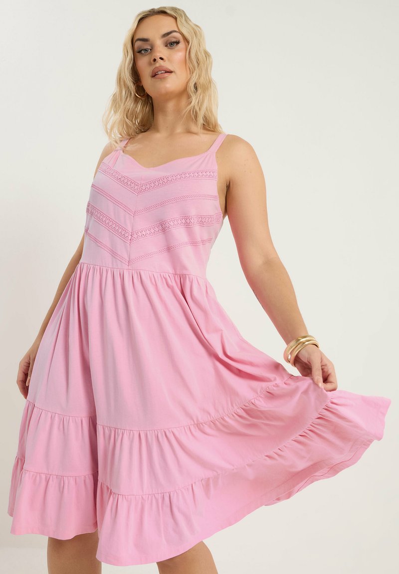 Mujer con un vestido rosa sin mangas de capas con detalles de encaje, sosteniendo ligeramente el vestido, complementada con pendientes de aro dorados y brazaletes.