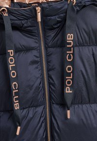 Marinblå pufferjacka med glänsande guldfoder, med en central dragkedja och svarta tygdragband präglade med "POLO CLUB" i guld.
