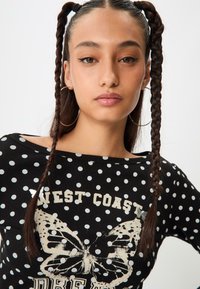 Čierny top s bodkami a off-shoulder strihom, s grafikou motýľa a textom "ZÁPADNÉ POBREŽIE." Štruktúrovaná tkanina a zapletené vlasy.