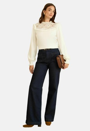 White long-sleeve top with a lace collar, paired with high-waisted dark blue wide-leg jeans. Brown clutch bag and heeled shoes complete the look.